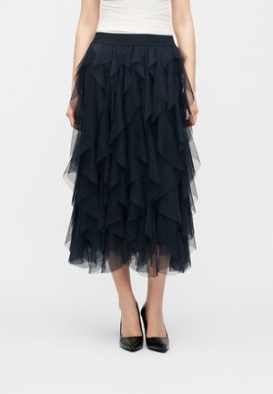 Jupe midi en tulle noir à plusieurs couches portée avec des talons hauts noirs et un haut blanc sans manches, ne montrant que le bas du torse et les jambes.