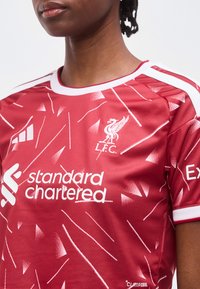 Κόκκινη φανέλα της Liverpool FC με λευκό γεωμετρικό σχέδιο, λογότυπα χορηγών και λευκή επένδυση στο γιακά και στα μανίκια, φορεμένη από άτομο.