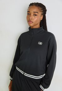 Schwarze Sportjacke mit hohem Kragen, Reißverschluss vorne und weißen Streifen an den Bündchen und dem Saum; verfügt über ein kleines Logo auf der Brust.
