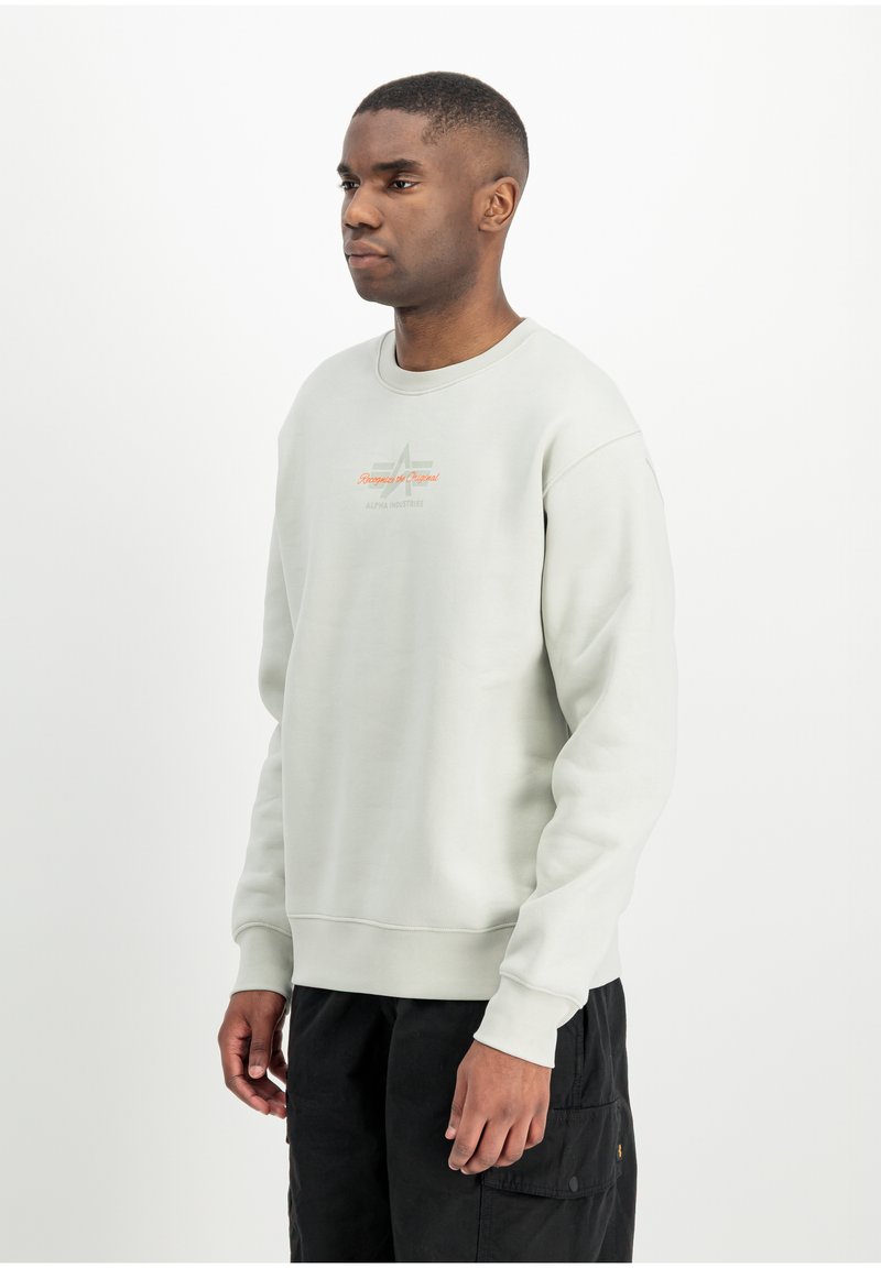 Jeune homme portant un sweatshirt gris clair avec un petit texte orange et un pantalon cargo noir, debout devant un fond blanc.
