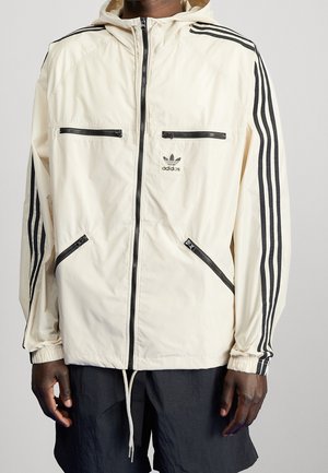 Jasno beżowa kurtka Adidas z czarnymi paskami na rękawach, zamkami błyskawicznymi z przodu oraz kapturem, noszona z czarnymi szortami.