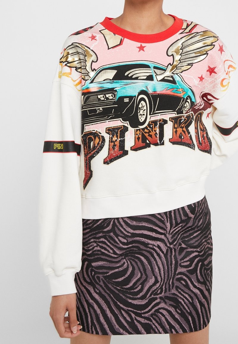 Personne portant un sweat blanc avec un motif graphique coloré de voiture ailée et le texte « PINKO », associé à une jupe imprimée zèbre noire et violette.