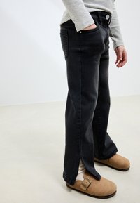 Schwarze Denim-Jeans mit lockerem Sitz, ausgestattet mit einem rohen Saum und Seitenschlitzen. Kombiniert mit hellbraunen Clogs und einem hellgrauen Langarmshirt.