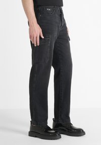 Pantalones cargo negros con un bolsillo lateral texturizado, corte recto y detalles de herrajes visibles. Están hechos de tejido de mezclilla. Footwear presenta un material negro brillante.