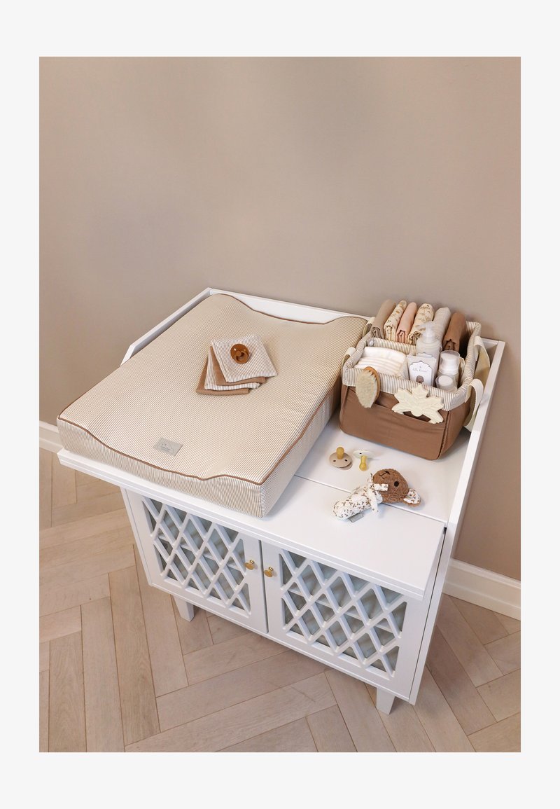 Μπεζ ριγέ μαξιλάρι αλλαγής μωρού σε λευκή ντουλάπα με οργανωμένο καλάθι που κρατά παιδικά ρούχα, μπουκάλια και παιχνίδια πάνω σε μπεζ τοίχο.