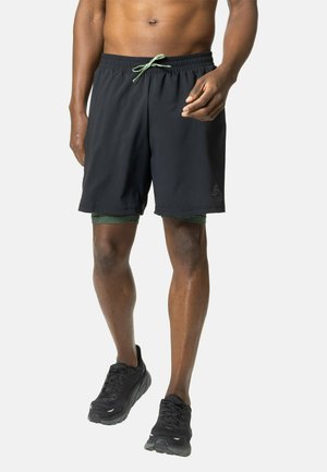 Black athletic shorts with a green drawstring, featuring a logo on the lower leg. Worn over dark green compression shorts and black sneakers.