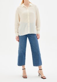 Femme portant une blouse crème transparente à boutons, un jean bleu à jambes larges coupé, et des sandales à talons hauts avec bride chevilles noires, se tenant sur un fond blanc.