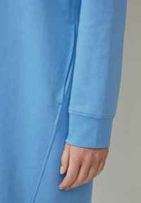 Hellblaues Sweatshirt mit glatter Textur, ausgestattet mit einer Seitentasche und einem Bündchenärmel. Der Stoff wirkt leicht und atmungsaktiv.