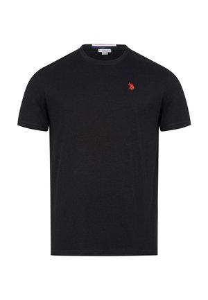 Mann geht und trägt ein schwarzes T-Shirt mit kleinem roten Logo, dunkle Jeans und weiße Sneakers vor einem schlichten hellen Hintergrund.