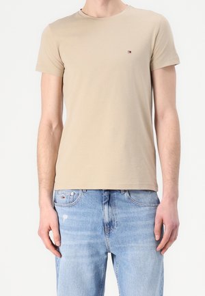 Beżowy dopasowany t-shirt z krótkim rękawem i małym logo flagi na piersi, noszony z jasnoniebieskimi, postaranymi dżinsami.