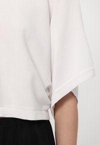 Valkoinen lyhyt sweatshirt, jossa on leveät hihat, teksturoitu kangas ja ribbireunus. Yhdistetty mustan vyötärönauhan kanssa, mikä tuo esiin löysän istuvuuden.