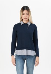 Dunkelblauer Strickpullover mit rundem Halsausschnitt über einem blau gestreiften Hemd, kombiniert mit hellblauen Jeans. Glatte Textur, schmaler Schnitt.