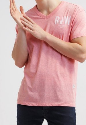 Personne portant un T-shirt rose à col en V avec l'inscription blanche "RAW", applaudissant, associée à un pantalon foncé sur un fond uni.