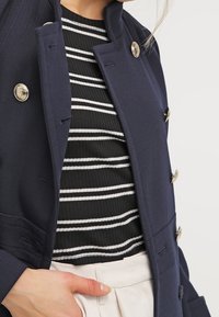 Cappotto doppiopetto blu navy con bottoni lucidi argentati, dotato di colletto strutturato e vestibilità sartoriale sopra una maglietta a righe bianco e nero.