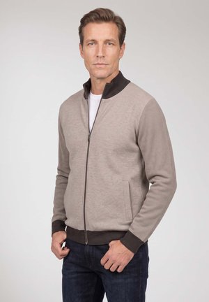 Mann stehend, trägt einen beigen Reißverschluss-Pullover mit dunkelbraunen Bündchen und Kragen über einem weißen Hemd und dunklen Jeans vor einem einfarbigen Hintergrund.