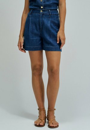Femme portant un short en jean bleu foncé taille haute et des sandales marron à brides, debout devant un fond uni.
