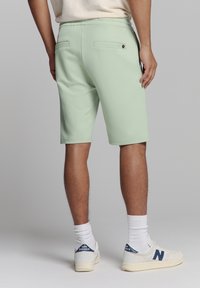 Lichtgroene shorts met een gladde textuur, voorzien van achterzakken en een klein logodetail. Gecombineerd met witte schoenen en enkelsokken.