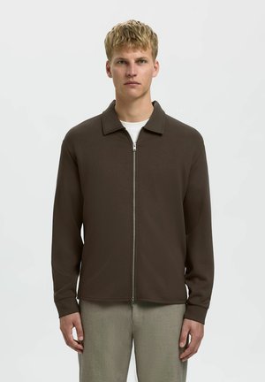Braune Zip-Jacke mit spitzem Kragen und langen Ärmeln, aus einem glatten Material gefertigt, über einem weißen Hemd getragen und mit hellen Hosen kombiniert.