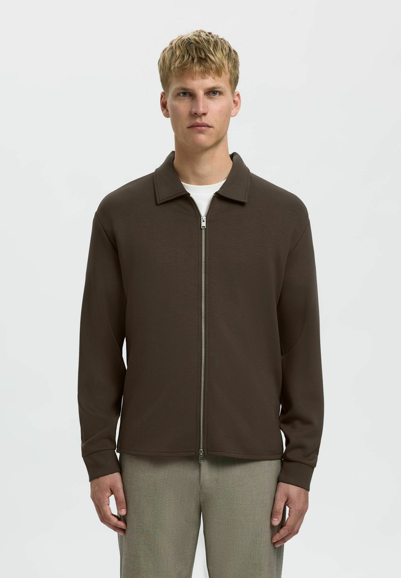Giacca marrone con zip, colletto a punta e maniche lunghe, realizzata in un materiale liscio, indossata sopra una camicia bianca e abbinata a pantaloni chiari.