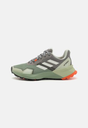 Zapatilla de trail running en gris y verde menta con parte superior de malla, acentos naranjas y suela texturizada. Cuenta con tres rayas blancas y un diseño acolchado.