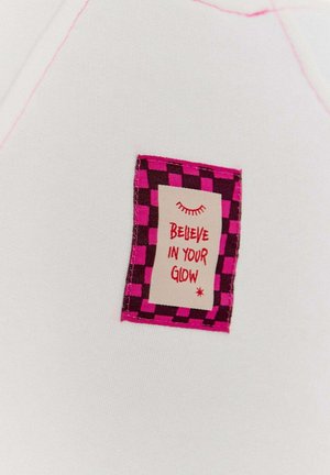 Biały materiał z różowym przeszyciem zdobi prostokątna łatka w różowo-ciemną kratkę, z napisem "WIERZ W SWÓJ BLASK".