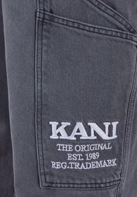 Šedá denimu látka s přišitým kapsou, na které je bíle vyšitý text: "KANI THE ORIGINAL EST. 1989 REG. TRADEMARK."