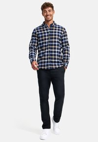 Camisa de cuadros abotonada en azul marino, crema y azul; cuenta con un collar clásico y mangas largas. Combinada con pantalones oscuros y zapatillas blancas.
