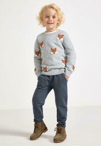 Lachend kind met krullend blond haar, gekleed in een grijze trui met oranje vosgezichten, blauwe broek en bruine veterschoenen, staande met de handen in de zakken.