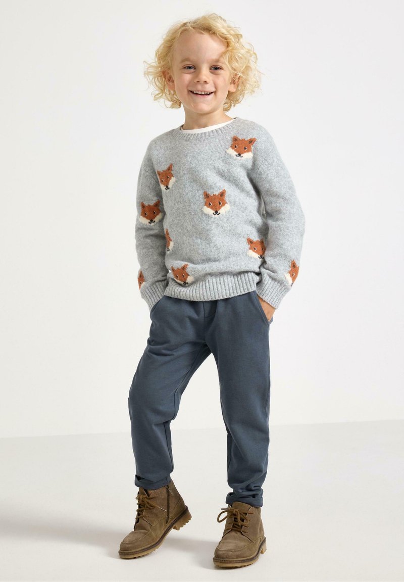 Lachend kind met krullend blond haar, gekleed in een grijze trui met oranje vosgezichten, blauwe broek en bruine veterschoenen, staande met de handen in de zakken.