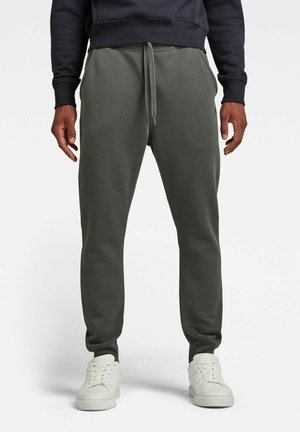 Mann trägt dunkelgraue Jogginghose mit Kordelzug und weißen Turnschuhen, steht vor einem schlichten weißen Hintergrund.