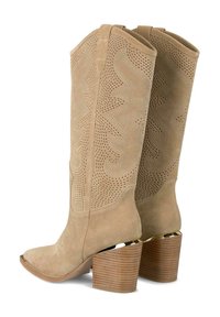Botas de cowboy de ante beige con punta afilada, recortes decorativos en el tubo y un tacón de madera grueso. Acentos en tono dorado en la base.