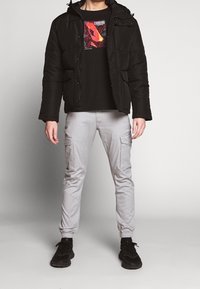 Zwarte gewatteerde jas met capuchon, zwart grafisch T-shirt met rood ontwerp, lichtgrijze cargobroek en zwarte sneakers.