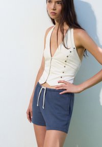 Top corto bianco senza maniche con scollatura a V profonda e bottoni laterali, abbinato a pantaloni corti di cotone blu con vita elastica e lacci.