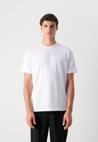 T-shirt en coton blanc avec un col rond et des manches courtes. Présente un logo brodé discret sur la poitrine. Associé à un pantalon noir.