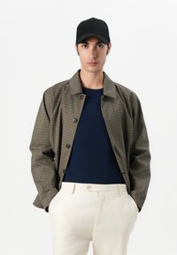 Checkered brown and black jacket with a lightweight design, worn over a navy top and cream trousers. Black cap completes the look.