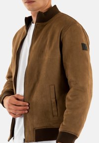 Giacca bomber in pelle scamosciata marrone con colletto a coste, chiusura zip e tasche laterali. Presenta un tessuto strutturato e un'etichetta con il marchio sulla manica.