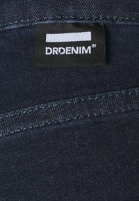 Tejido de mezclilla oscura con costuras visibles, que presenta una etiqueta negra con texto blanco que dice "DRDENIM". Textura suave y forma estándar.
