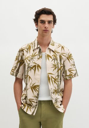 Junger Mann trägt ein beigefarbenes kurzärmeliges Hemd mit grünem Bambusmuster über einem weißen Tanktop und olivgrünen Hosen, Hände in den Taschen, neutraler Hintergrund.