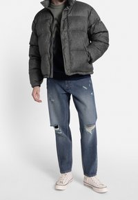 Uomo che indossa un giubbotto imbottito grigio scuro, un maglione scuro a fantasia, jeans blu strappati e sneakers alte bianche in piedi contro uno sfondo neutro.
