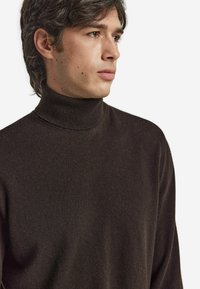 Młody mężczyzna o brązowych włosach, ubrany w ciemnobrązowy sweter z golfem, patrzący w bok na tle jednolitego białego tła.