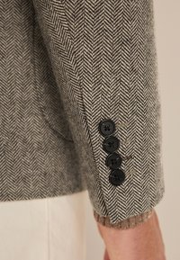 Grau gemusterte Blazer-Ärmel im Fischgrätenmuster mit vier schwarzen Knöpfen und einem gestrickten Bündchen, das die gewebte Textur und das elegante Design zeigt.