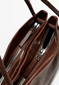 Brown leather handbag interior featuring two compartments, silver zippers, and textured lining. Smooth finish with detailed stitching.
