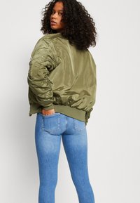 Olivgrön bomberjacka med glänsande, slät yta, ribbade ärmslut och nederkant. Bärs över ljusblå, figurnära jeans, och visar en bakvy.
