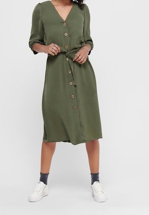 Woman wearing a knee-length olive green button-front dress with 3/4 sleeves, tied belt, gray socks, and white sneakers standing against white background.