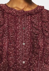 Blusa bordeaux con colletto arricciato, tessuto testurizzato con motivo floreale e bottoni argentati lungo la patta frontale.