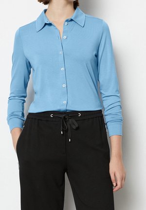 Frau trägt ein hellblaues, langärmliges Hemd mit Knöpfen, das in eine schwarze Hose mit Kordelzug gesteckt ist, die Hände in den Taschen.