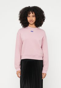 Sudadera rosa de cuello redondo, mezcla de algodón, con puños y dobladillo acanalados, pequeño parche de logo azul, combinada con una falda negra plisada.