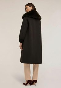 Lång svart kappa med en pälsig krage, vida ärmar och sidofickor, tillsammans med ljusa skräddarsydda byxor och burgundyklackar. Slät textur.