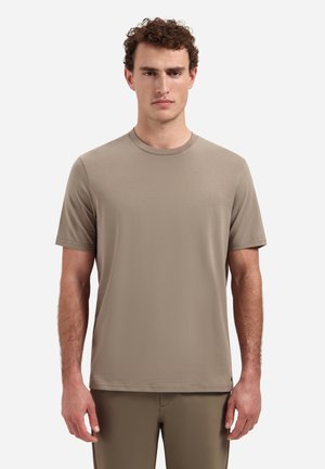 Man met krullend haar, gekleed in een effen beige T-shirt met korte mouwen en bijpassende broek, staand tegen een witte achtergrond.