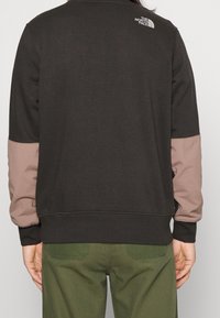 Svart sweatshirt med ljusbruna detaljer på ärmar, ribbade muddar och nederkant. Har en vit logotyp på baksidan av nacklinjen.